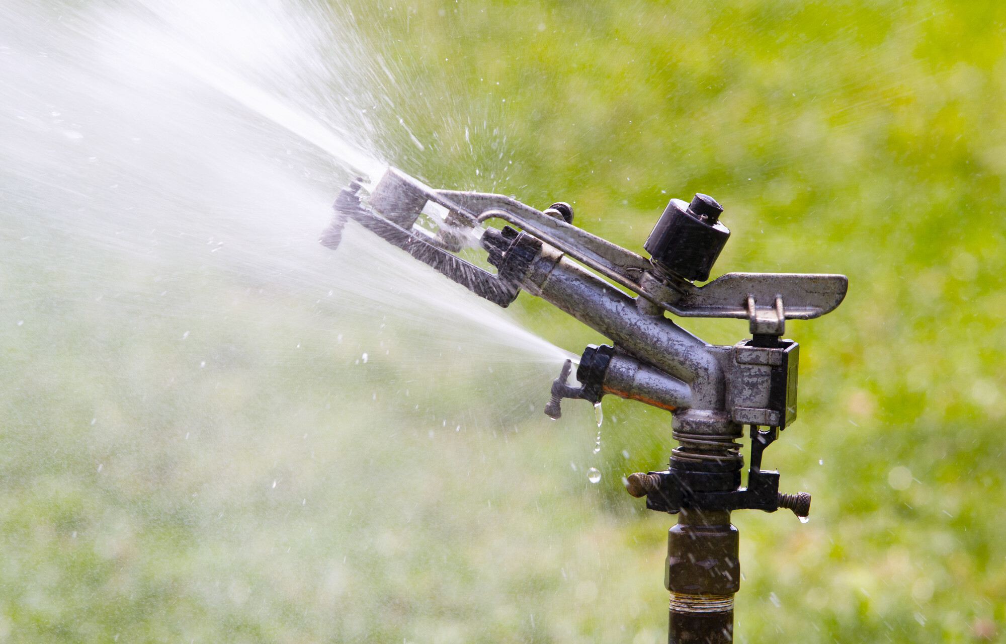 Irrigatore a impulsi in metallo su asta che spruzza un getto d'acqua sopra un prato verde sfocato